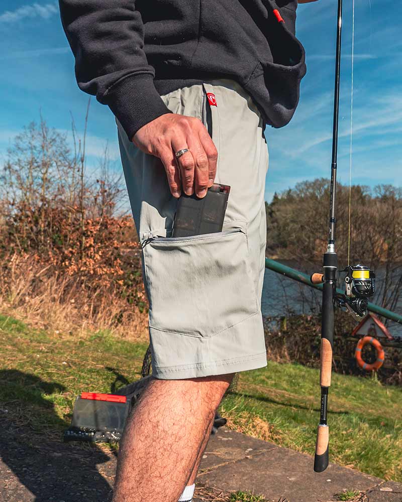 Hombre usando pantalones cortos ligeros Fox Rage en gris, con bolsillo y caña de pescar al fondo.