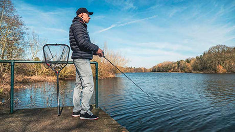 Hombre pescando en un lago con pantalón desmontable Fox Rage, rodeado de naturaleza y cielo despejado.